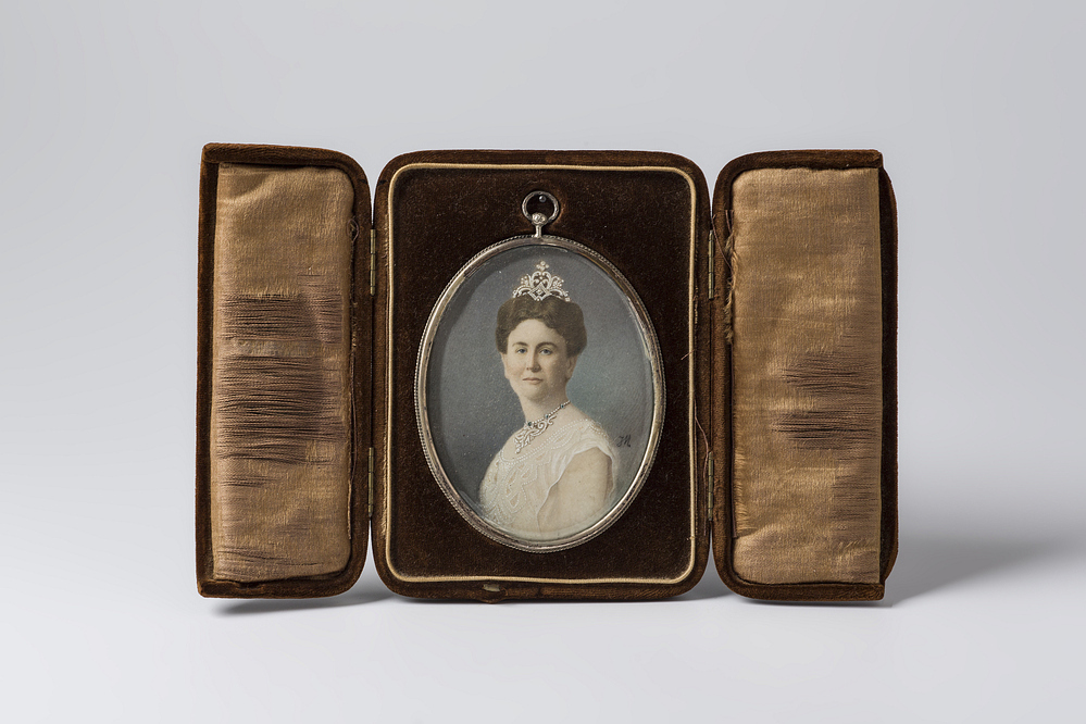 Ovaal portret van Koningin Wilhelmina, in een uitklapbaar reisdoosje.  Portret : busteportret van de koningin in een lichte japon, op zachtblauwe achtergrond. R.o. de initialen J H.  In zilveren lijstje met haakje,  met op de achterzijde de inscriptie : "Wilhelmina Helena Paulina Maria Koningin der Nederlanden 6 Sept. 1898 - 6 Sept. 1923. "   Hieronder een merkje : een M, en daaronder drie puntjes. Het portret rust in een met bruin velours afgewerkt doosje, waarvan het deksel in twee helften uitgeklapt kan worden. Aan de achterzijde zit een uitklapsteun, zodat van het geheel een  drieluik-foto opstelling gemaakt kan worden.