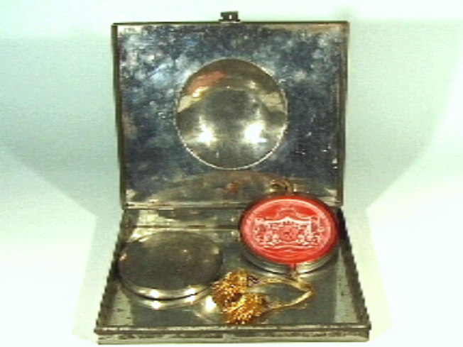 Blikken platte trommel, met in deksel een bolle ronde uitstulping. Hierin een rond zegel in rode was met afbeelding van  Wilhelmina