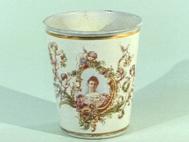 Geëmailleerde blikken beker, met op witte ondergrond portret van jonge koningin Wilhelmina, engeltjes, bloemen en groen, kroon, koningswapen.