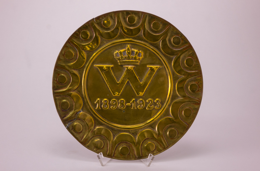 Bliken bord, verguld, met op het plat een hoofdletter W, een kroon, en "1898-1923".
