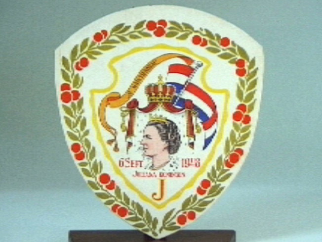 Kartonnen schildje  met op witte ondergrond een kleurige afbeelding met o.a. portret Juliana, kroon, vlag, en rand van oranjeappeltjes.