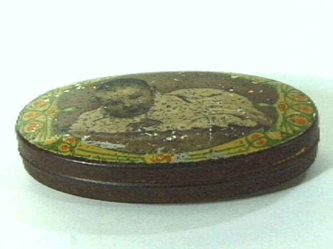 Ovaal blikken doosje met deksel, waarop een afbeelding van Juliana als baby liggend op de buik, omringd door decoratie in groen/oranje/geel.