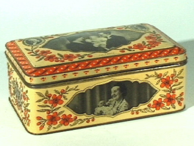 Rechthoekige koektrommel van blik met deksel, in crème, oranje, goud, met afbeeldingen in zwart-wit.