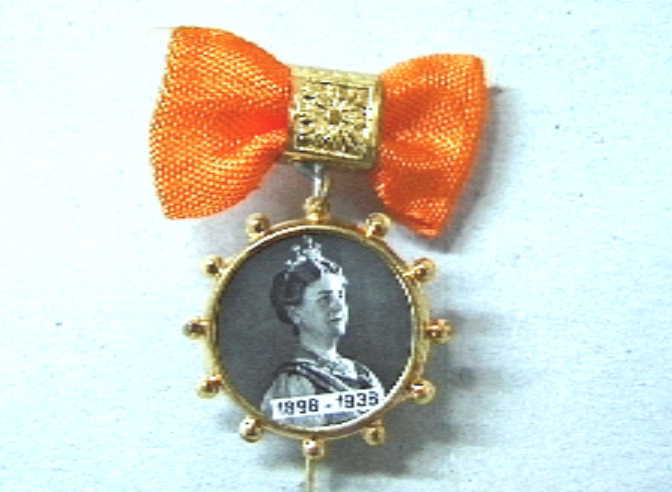 Steekspeldje met hangend aan een oogje een rond miniatuur fotoportret van koningin Wilhelmina en het opschrift 1898-1938.