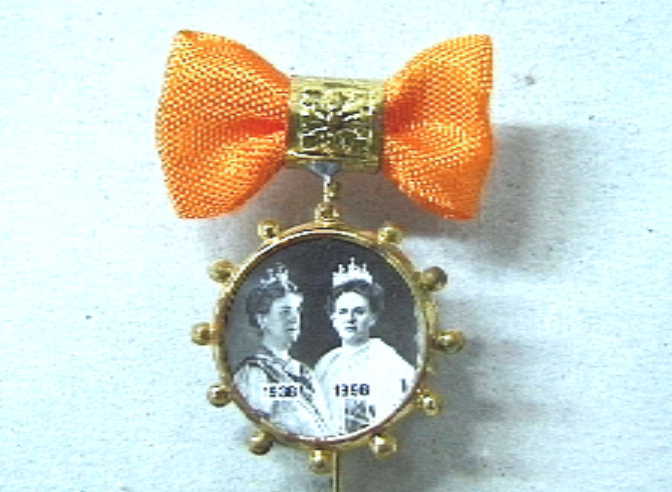 Steekspeldje met hangend aan een oogje een rond fotoportretje met twee busteportretten van koningin Wilhelmina uit 1938 en 1898.