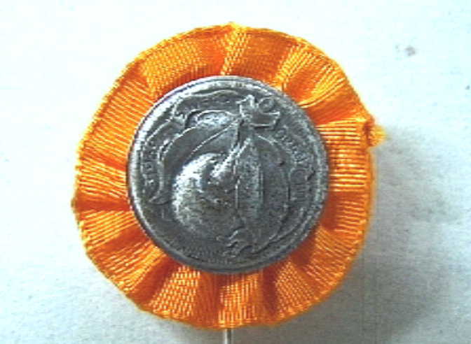 Rond speldje met afbeelding van een ronde vrucht (oranjeappeltje) met twee grote bladeren.