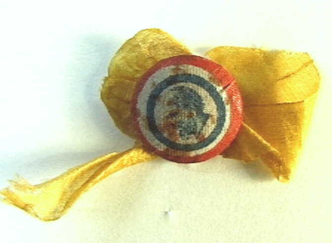 Corsage, bestaande uit oranje lintje en een ronde knoop met op textiel een portret naar links van koning Willem III in een rood-wit-blauwe omlijsting.