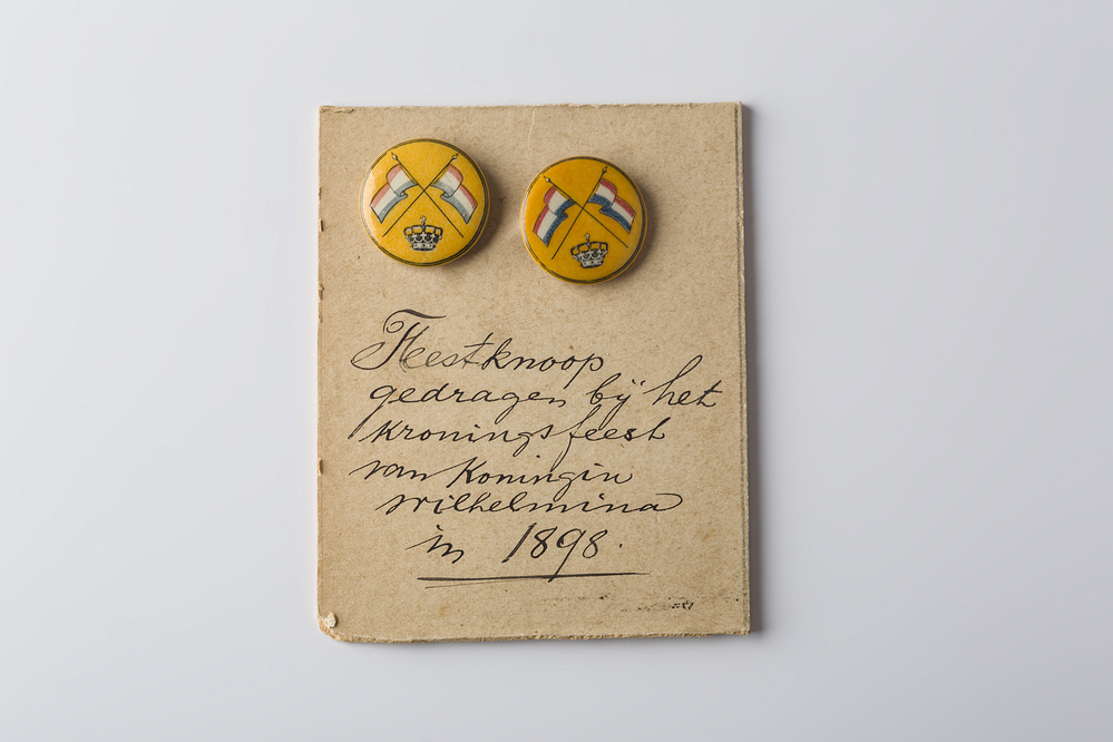 Twee feestknopen, oranje, met decoratie van gekruiste vlaggen en een kroon.