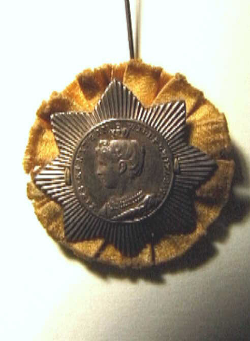 Corsage, stervormige beeltenis van de jonge koningin op oranje stof. In een rond doosje met feestopdruk.  J.M. van Kempen en zonen.