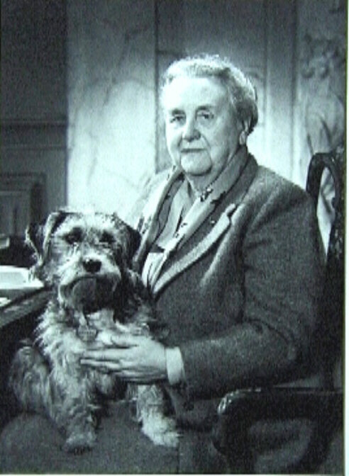 Foto van Wilhelmina op oudere leeftijd, zittend in leunstoel naar links, met hond Blackie op schoot. Kleding ; wollen mantelpak, sjaaltje.