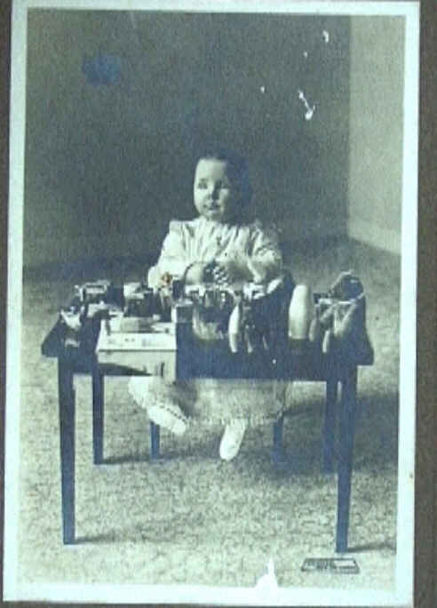 Foto van Juliana als peutertje, zittend achter een tafeltje met speelgoed.