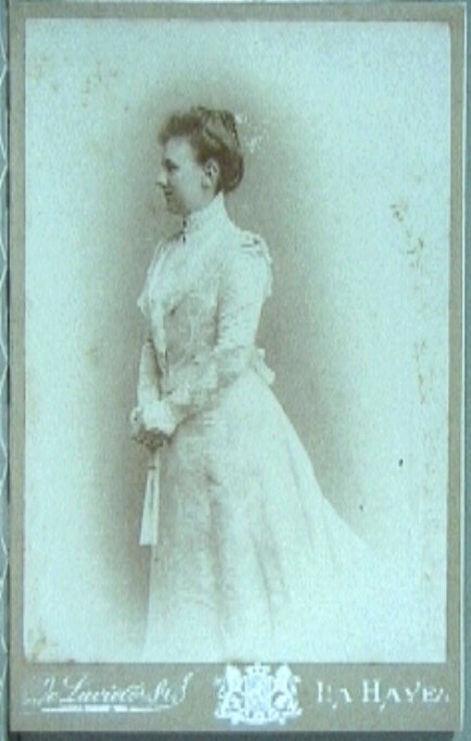 Portret van Wilhelmina , staand naar links, tot aan haar knieen, in witte japon met waaier in haar voor haar buik gevouwen handen.