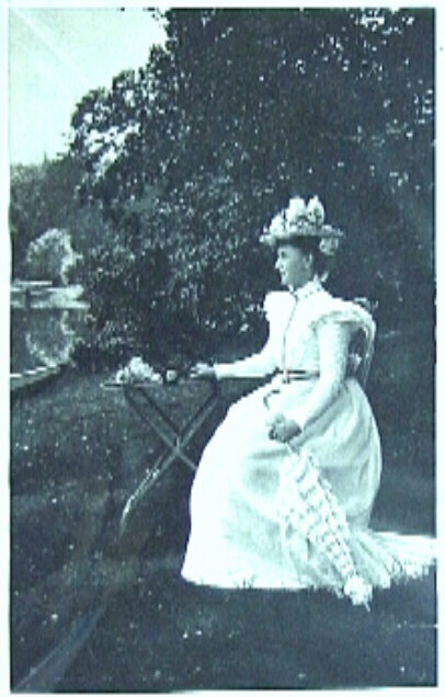 Wilhelmina met lichte jurk, hoed en parasol, zittend aan een tafeltje met daarop rozen, in het park van het Paleis te Soestdijk.
