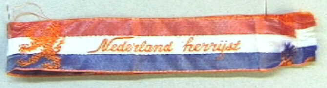 Lint in de kleuren rood-wit-blauw, met in oranje ingeweven de Nederlandse Leeuw, en het opschrift "Nederland herrijst".