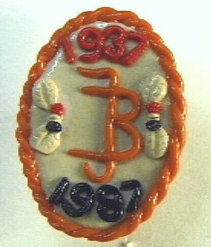 Ovale corsage met speld aan de achterzijde. Op crème ondergrond met oranje omlijsting de verstrengelde initialen J B, boven in rood 1937, onder in blauw 1987.