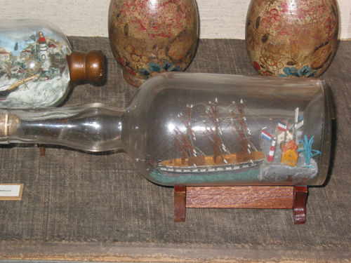 Miniatuur volschip in fles met op de achtergrond twee huisjes en een molen.