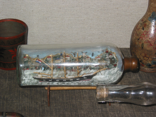 Driemastvolschip "Tasman" met Nederlandse vlag en zwarte romp in fles op houten staander aan de kust met huisjes, molen, vuurtoren aan de haven.