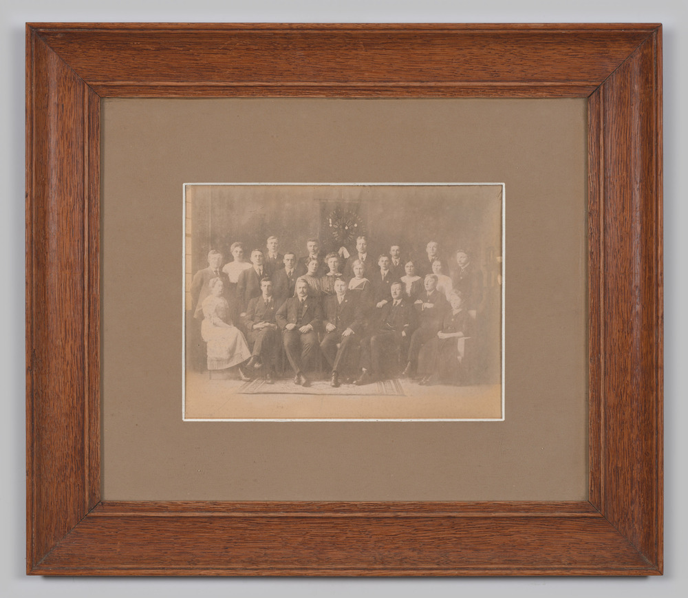 Foto in lijst, achter glas, voorstellende groepsportret van Oefening en Volharding