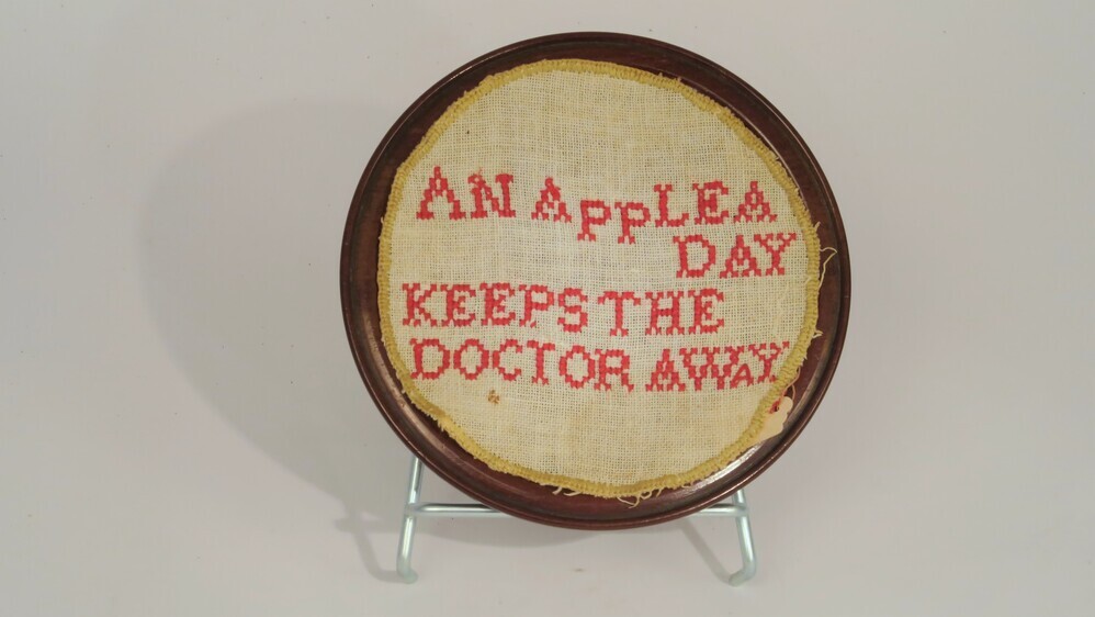 Rond houten schaaltje, voorzien van bijpassend borduurwerkje met de tekst "AN AppLE A DAY KEEPS THE DOCTOR AWAY"