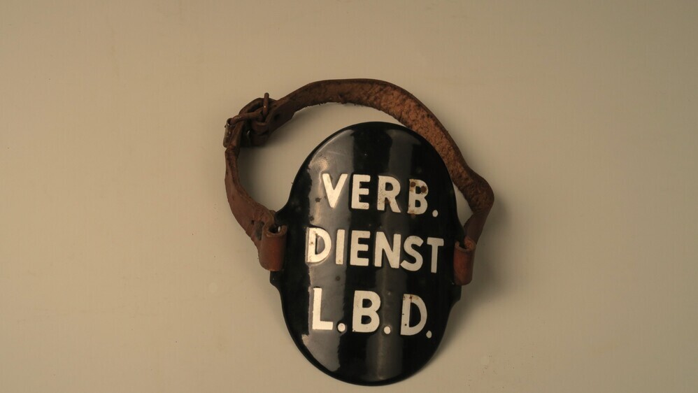 Geëmailleerd, metalen armplaat ('schildje') dat met twee leren riemen aan de arm kan worden gegespt om te worden gedragen met de opdruk Verb. dienst (Verbindingsdienst) L.B.D. (luchtbeschermingsdienst)