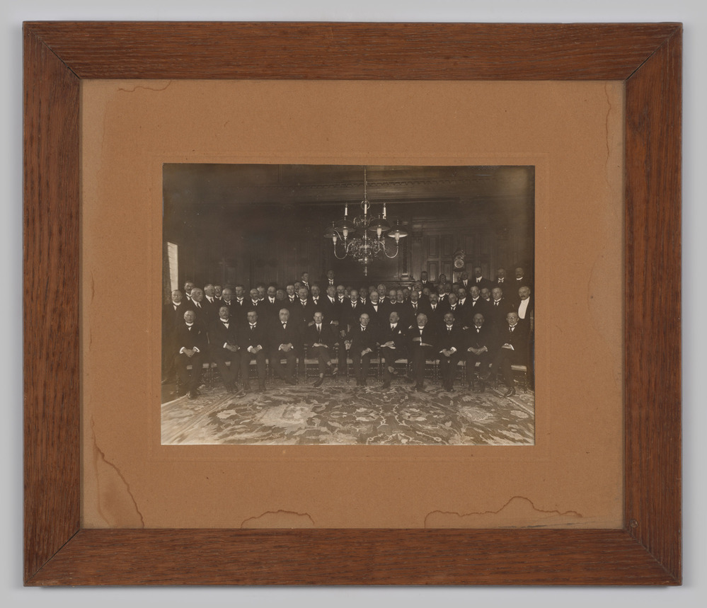 Foto van een grote groep heren in deftig interieur, ingelijst, achter glas