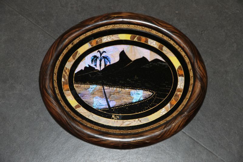 ovale glasschildering van een gezicht op de baai te Rio de janeiro, met op de achtergrond de rots met het kruis, alles in het zwart en goud, opgevuld met vlindervleugels, rand eromheen vlindervleugels, daarna rijkelijk bewerkte houten rand en uiteindelijk houten sierlijst.