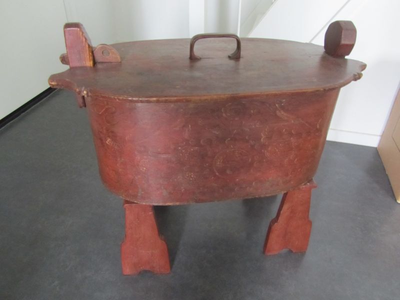 houten ovalen bruine butje, aan de beide uiteinden van de kuip zit aan de binnen- of de buitenzijde een verticaal houtje, dat 'n eindje boven de rand uitsteekt en een verdikt of beneden kopje heeft. Het deksel nu heeft aan beide einden een paar uitbuigingen waartussen een opening wordt gevormd. Door nu gebruik te maken van de buigzaamheid van het materiaal kan men het deksel eerst aan een kant onder de verbrede kop schuiven en daarna „snapt" het andere uiteinde om het tweede „kopje". Deze grote butje heeft een ijzeren pen om het deksel te fixeren op haar plek, zodat het deksel niet zomaar te verwijderen is en gesloten blijft. De zijkant is gedecoreerd met bloemmotieven en een bloemenvaas. Het geheel staat op 2 losse steuntjes. De decoratie is 18 de eeuw.