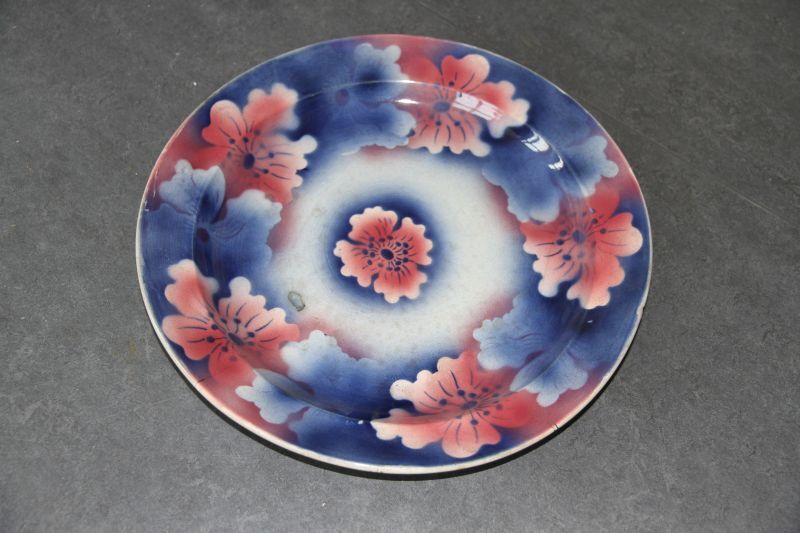 grote bord in blauw en wit met op de rand roze oranje en blauwe bloemen, in het midden enkele roze oranje bloem, aan achterzijde stempel KuznetSov Budy fabrieksmerk 1890 - 1917