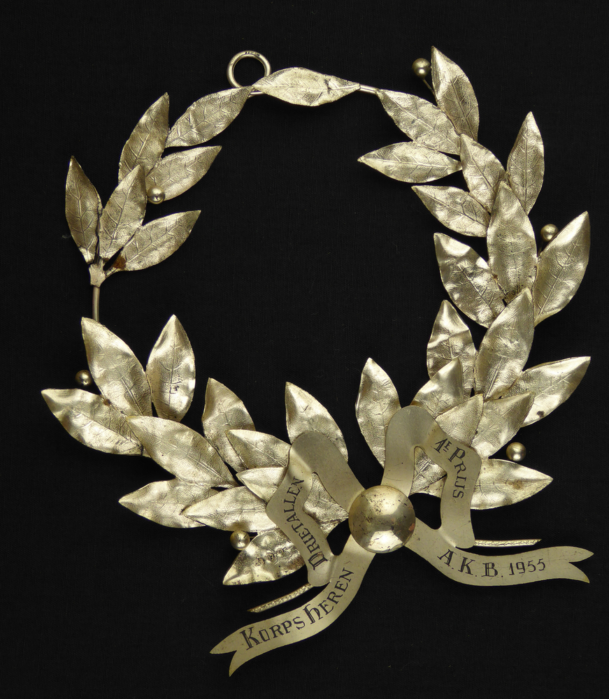 sportprijs in de vorm van een lauwertak met strik , aan onderkant gegraveerde inscriptie korps heren drietallen 1e prijs A.K.B. 1955, op blad gehalteteken zilveren leeuw, minervahoofd jaarletter G = 1941 en 10 meesterteken JC = Fa J & J.P. Crusio Arnhem