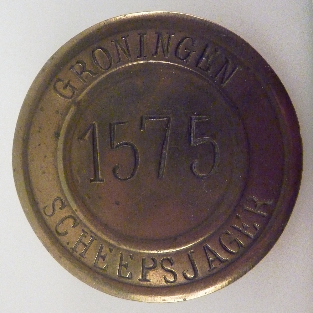 ronde messing Scheepsjagerspenning met nummer 1575 van Hendrik Brul te Nieuweschans. Bij deze penning is de originele bijbehorende akte bewaard gebleven en bevind zich ook in de colllectie van het Veenkoloniaal Museum.