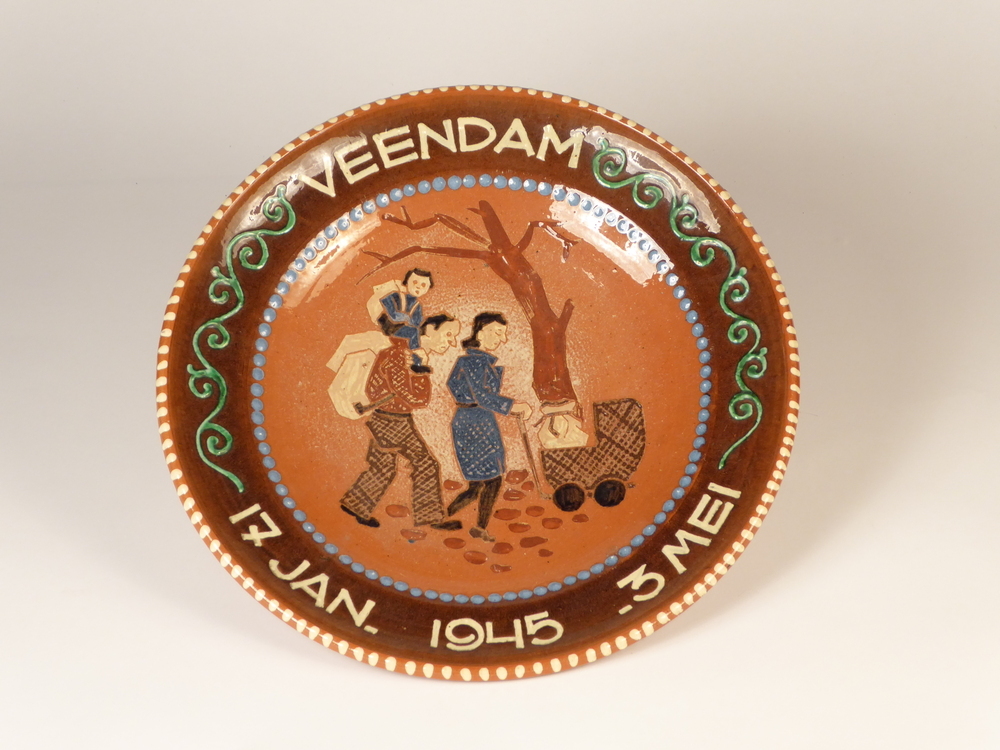 Een ronde grote uit roodbakkende aardewerk bestaande sierbord met binnenin een afbeelding van een vluchtende gezin, bestaand uit een man, vrouw met kind en kinderwagen hobbelend over de kasseien en op de achtergrond een eenzame boom, wat symbool staat voor de verwoesting van Nederland. Op de rand in witte belettering Veendam 17 Jan 1945 – 3 mei. Een gedenkbord ter herinnering aan de in Veendam ondergebrachte evacues uit Venlo.