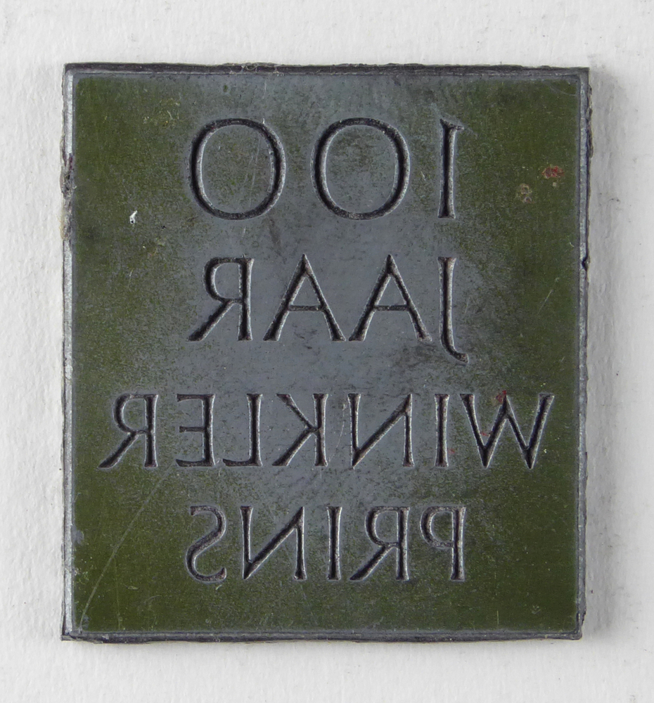 vierkante non ferro metalen cliche met inscriptie 100 jaar Winkler Prins, afkomstig van kantoorinstallaties Bv Schuur te Veendam, achterkant blauw