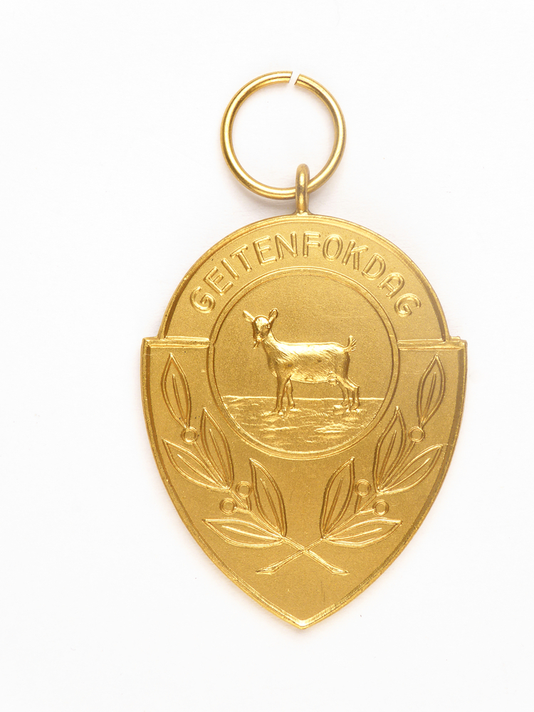 goudkleurige medaille in de vorm van een schild met ronde bovenkant, afbeelding van een geit en bladeren en inscriptie geitenfokdag, op achterkant inscriptie prov k 7-9-57 1E pr 3 j A 103 H Dijkstra