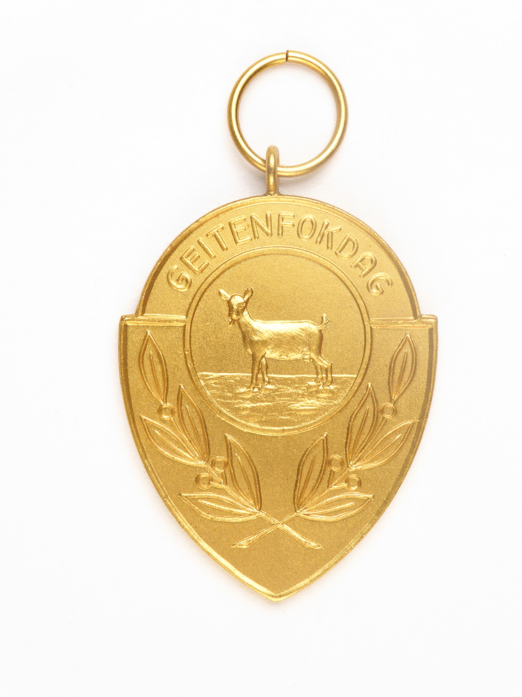goudkleurige medaille in de vorm van een schild met ronde bovenkant, met afbeelding van een geit en bladeren en inscriptie geitenfokdag, met op achterkant inscriptie prov k 7-9-57 1A pr Coll Afd veendam