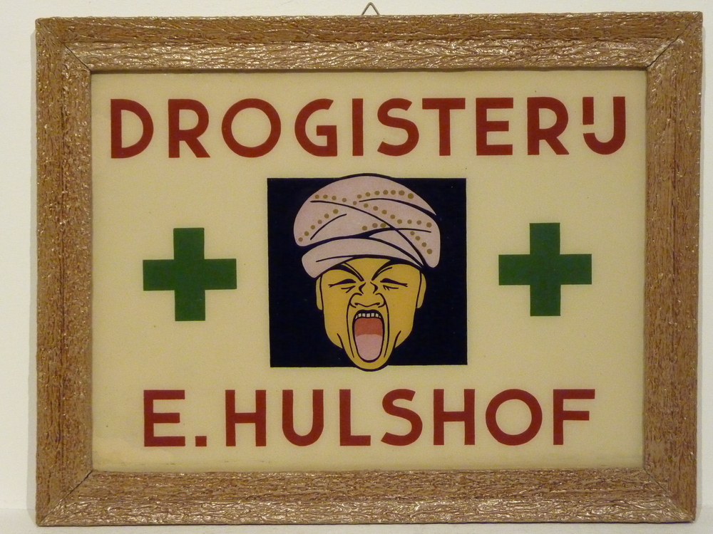 kartonnen bord in hout met gipsen lijst met opdruk drogisterij E Hulshof, afbeedling gezicht met open mond, weerszijden een groene plusteken