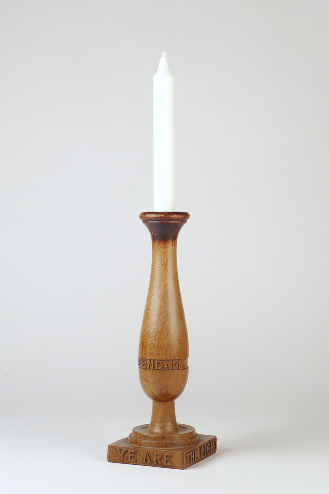 smalle hoge balstervormige kandelaar op voetstuk met bovenin geplaatste rood koperen kaarsenhouder, aan voorkant buik ingesneden inscriptie Veendam 1945, rondom voetstuk gesneden inscriptie :  Ye are the light of the World St M 5 - 14 – 15 (het getal 15 is helaas beschadigd en geheel verdwenen) = Mattheus 5 vers 14 , 15. In de bijbel staat hier : Mattheüs 5 : 14 Jullie zijn het licht in de wereld. Een stad die boven op een berg ligt, kan niet verborgen blijven. 15 Men steekt ook geen lamp aan om hem vervolgens onder een korenmaat weg te zetten, nee, men zet hem op een standaard, zodat hij licht geeft voor ieder die in huis is. In opdracht gesneden door beeldhouwer Hendrik Anthonius ter Reegen uit Veendam en geschonken door mevrouw M Ter Borg uit Holten. Een zelfde soort kandelaar gaven de ouders van mevr. Ter Borg (toen wonende te Veendam) mee aan Lorne Baldassero, Canadese officier van het bevrijdingsleger die 7 maanden bij hen ingekwartierd waren.