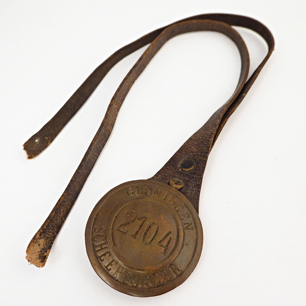 ronde messing scheepsjagerspenning compleet met leren riem, met op voorkant geslagen inscriptie Groningen 2104 Scheepsjager. Scheepsjagerspenning afkomstig van Barteld Woortman, Nieuwediep, werd met 35 jaar scheepsjager