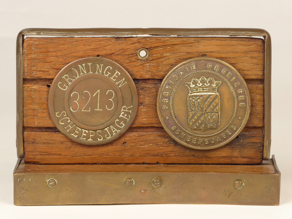 twee op hout gemonteerde scheepsjagerspenningen met messing rand omvat en aan achterkant metalen steun, een met randschrift Provincie Groningen scheepsjager rond provinciewapen de ander met randschrift Groningen Scheepsjager rond nummer 3213 het laatste exemplaar sinds 22 oktober 1936 gedragen door Jozef Prins te Nieuwe Pekela.