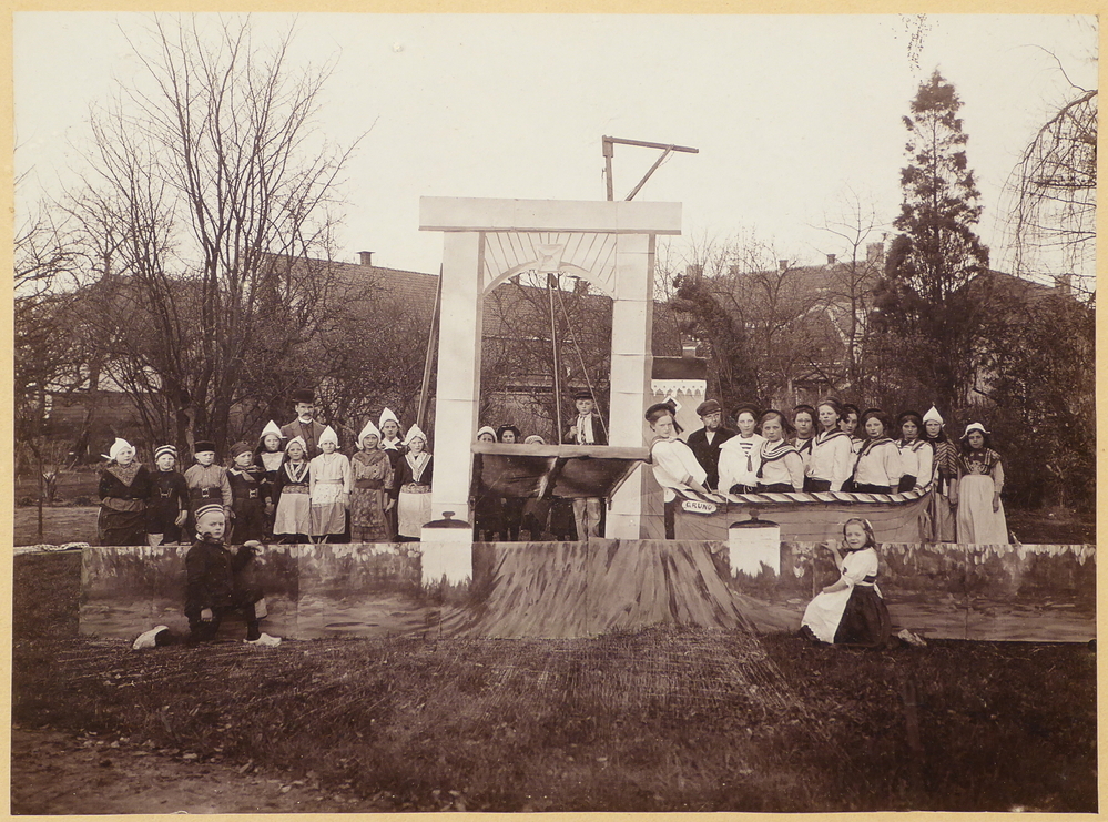 Ingelijste foto in een goudkleurige gipsen lijst met hier en daar beschadigingen aan de zijkant. De achterkant zit los. Het geheel is 40 x 35 cm, de foto slechts 23 x 17 cm. Op de foto poseren een aantal kinderen in klederdracht met een witte floddermuts op naast een klapbrug, aan de rechterkant een aantal kinderen in matrozenpakjes in een boot met de naam Gruno. Links en rechts van de klapbrug knielen een jongen en meisje terwijl ze een geschilderde kademuur vasthouden. Achter de kinderen aan de linkerkant staat een man met een dikke snor en hoed. De foto is genomen in een tuin, en op de achtergrond zie je nog enkele huizen. De fotograaf is H. Loman van het Beneden Westerdiep, getuige een stikker rechtsonder. Op de achterkant is met potlood geschreven: K. Grasmeijer Sappemeer Slochterstraat. Als je de achterkant verwijderd leest je achterop de originele foto met pen: Mijn vader met een gedeelte van Wildervankster Zangklassen waarvan hij toen directeur was achter het voormalige Hotel “De Nederlanden” toen in bezit van Benno Smit. Opname nog voor 1922. B.H.J. ( Bregine Henriëtte Imkéa ) uit den Boogaart – Grasmeijer 26-07-1989. De man waarom het hier op de foto gaat is Keimpe Grasmeijer. Hij werd geboren op 4 mei 1881 te Sappemeer als zoon van dagloner / fabrieksarbeider Geert Grasmeijer en Imke Munneke. Hij huwde 14 mei 1910 te Sappemeer met Hendrika van der Bend. Hij was achtereenvolgens onderwijzer  te Kleinemeer (1901), onderwijzer aan de Zuiderschool te Wildervank (1910), hoofd van de school te Eexterveenschekanaal (1915), hoofd van de school te Wildervanksterdallen (1922) en hoofd van de Westerschool te Wildervank (1931). Hij overleed op 27 april 1937 te Wildervank. Met zijn schoolkinderen gaf hij uitvoeringen, operettes en concerten. Tussen 1913 en 1914 traden de zangklassen en jongedamesafdelingen meerdere malen op in de tuin van hotel de “Nederlanden” met een diverse programma. Een van die programma’s was “Wachter Ahoi”, waarop men op het toneel achter een echte wal een bootje zag varen. Fotograaf Loman legde de hoofdrolspelers voor dit toneelstuk getuige deze afbeelding van tevoren nog even vast. Het geheel werd tijdens de voorstelling muzikaal bijgestaan door de heer Van Halteren op piano en B. Smit hielp met de decoraties. Een kinderuitvoering heeft altijd een bijzondere aantrekkelijkheid, want de ouders genieten blijkbaar al van de aanblik van de door geestdrift gloeiende kindergezichtjes. De kinderen in hun luxueuze kostuums, verlicht door de talrijke elektrische lampjes geeft een aardig effect.