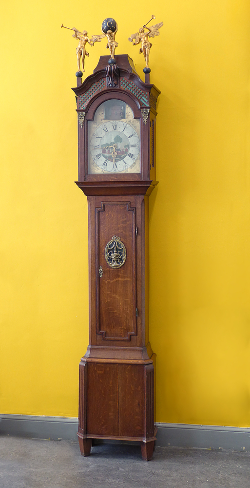 Een staand horloge in eiken kast. De dubbele kap met gecontourneerde bekroning met 2 verguld houten bazuin blazende engelen en een dito Atlas. Voorzien van zaagwerk. Polychroom gedecoreerde wijzer plaat met in de toog een wachtlopende c.q. wandelende soldaat. De wijzerplaatring met Romeinse en Arabische cijfers geeft de uren en de minuten aan. Adres: ‘Jan J Bruins’ en ‘Willem Holle’ en datering ‘1799’. Het uurwerk met gaand- en slagwerk op het halve en het hele uur. Hoogte (exclusief de kapbekroning) 225 cm.
