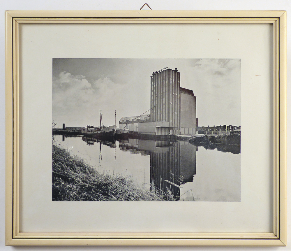 foto in witte houten lijst met daarop een pakhuis van de COBO ( Cooperatieve Aan- en Verkoopvereniging Boerenbelang ( opgericht in 1940 als Boerenbelang G.A. te Sappemeer, in 1965 door het samengaan van Coöperatie Noord-Oost te Veendam en Coöperatie Boerenbelang te Sappemeer gefuseeerd tot COBO, fuseerde met de aankoopcentrale in 1983 tot ACECO bv, fuseerde weer in 1990 tot ACM en in 2010 tot AGRIFIRM) te Hoogezand aan de van neckstraat. In het kanaal liggen 2 coasters te wachten, de achterste is coaster Tasman.