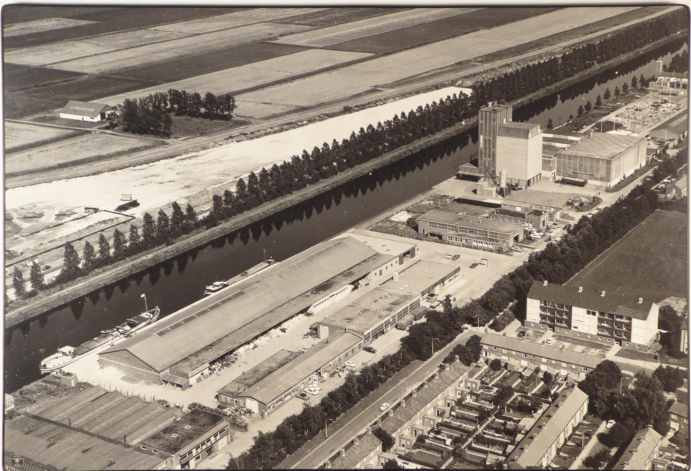 grote luchtfoto op MDF plaat met zwarte kunststoffen rand en 2 metalen beugels aan achterzijde voor de ophanging met daarop het pakhuis van de COBO ( Cooperatieve Aan- en Verkoopvereniging Boerenbelang( opgericht in 1940 als Boerenbelang G.A. te Sappemeer, in 1965 door het samengaan van Coöperatie Noord-Oost te Veendam en Coöperatie Boerenbelang te Sappemeer gefuseeerd tot COBO, fuseerde met de aankoopcentrale in 1983 tot ACECO bv, fuseerde weer in 1990 tot ACM en in 2010 tot AGRIFIRM) te Hoogezand aan de van neckstraat, genomen op 13 mei 1969. In het kanaal liggen verderop 2 schepen te wachten om te woren geladen of gelost.