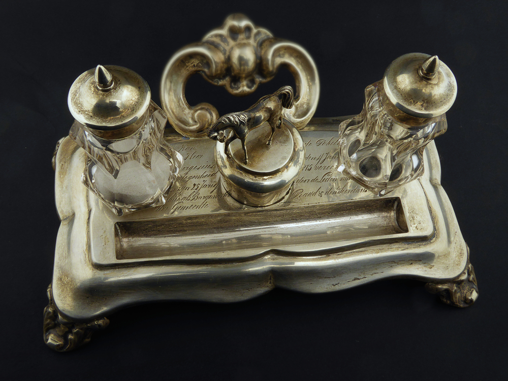 Zilveren rechthoekig inktstel staand op 4 omgekeerde acantusblad vormige pootjes met 2 kristallen geslepen zandstrooiers met zilveren puntdeksel en lakzegelwashouder met op deksel een paard, aan achterkant een opengewerkte handgreep, met aan bovenkant inscriptie : aan de heer T. Borgesius bij gelegenheid van zijn 25 jarig jubilé als burgemeester der gemeente Oude Pekela op den 4 januari 1875 vereerd door de Leden van de Raad & den Secretaris. Aan achterkant gemerkt gehalteteken leeuw en letter U = 1879, deksel lakzegelwashouder gemerkt K 1 in zeshoek , zilvermerk gebroeders Hendrik Geerts Kanning & Jan Geerts Kanning, zilversmeden te Leeuwarden 1866 – 1868. Is dus eerder gemaakt en later pas gegraveerd.