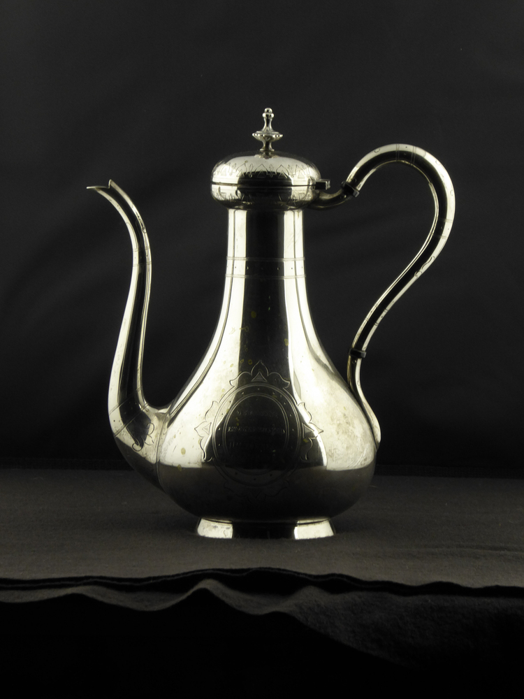 Ranke hoge zilveren koffiepot op standring, breedte van tuit tot oor 22,5 cm, hoogte 25,5 cm, slanke tuit met krulmotiefjes, ranke gebogen oor met decoratieve krulmotiefjes bestaat uit 3 delen met zwarte kunststoffen tussenringen, geheel boven en onder vastgezet met 2 zilveren pennetjes, scharnierende iets bolvormige deksel met rondom geometrische motieven, dikke rechthoekige scharnier, klein gekartelde bloemornamentje met punt bovenop, aan binnenkant deksel gemerkt met minervahoofd, aan onderkant binnenkant standring gemerkt met jaartalletter S = 1877 en erg versleten zilversmidsmerk A halve K met daaronder iets in de vorm van een driehoek, restant van een cijfer = waarschijnlijk AK 47 = Adrianus Kuijlenburg PZ te Schoonhoven, 1862 – 1882, of AK 49 = Albert Kuipers te Workum, 1871 - 1925, lopende leeuwtje 2 = 2e gehalte zilver voor klein zilveren werk, aan voorkant in gecontourneerde ovaal schild met een geornamenteerde omlijsting gegraveerd: “geschenk van broeders en zusters aan N.H.Tonkes & aan G.A.Fiebens “, aan achterzijde in gecontourneerde ovalen schild met een geornamenteerde omlijsting gegraveerd:  “1853 huwelijksvereeniging 1878 ”. 
Nanno Hommes TONKES, geboren Meeden 19-10-1820, overleden Heereweg (gem. Meeden) 01-07-1892, zoon van Hommo Edzes TONKES en Wiepke Nannes SIERTS.
Hij is getrouwd (1) te Meeden 11-08-1842 met Zwaantje Luitjens BOUWMAN, geboren Meeden 08-04-1818, overleden aldaar 05-08-1845, dochter van Luitje Luitjens BOUWMAN en Eetje Menzes TONKES. Uit dit huwelijk:
1.Luitjen Nannes TONKES, geboren Meeden 26-05-1843, overleden Solwerd 14-09-1908. Landeigenaar (1895). Hij is getrouwd Appingedam 22-03-1895 met Hilje TOXOPEUS, geboren Opwierde 13-11-1859, overleden Solwerd 10-01-1939, dochter van Tjark Geerts TOXOPEUS (houthandelaar) en Anje Luurts HOEKSEMA. Luitjen hertrouwt Appingedam 06-06-1867 met Catharina BONTHUIS TICHELAAR, geboren Tjamsweer 30-09-1842, overleden Solwerd 14-01-1889, dochter van Derk BONTHUIS TICHELAAR (landgebruiker) en Jantje Hindriks de BOER.
2.Hommo TONKES, geboren Meeden 05-06-1845, overleden Meeden 27-08-1845.
Nanno hertrouwt (2)Meeden 28-01-1853 met Geertruida Anna FIEBENS, geboren Westerbroek, overleden Heereweg (gem. Meeden) 02-05-1909, dochter van Luitje Geerts FIEBENS en Margien EEFTING. Uit dit huwelijk:
3. Lucia Maria TONKES, geboren Meeden 05-01-1859, overleden Heereweg (gem. Meeden) 09-09-1910. Zij is getrouwd Meeden 09-06-1881 met Roelof DOJES, geboren Uithuizen 23-09-1854, overleden Groningen 14-05-1930, zoon van Renger Roelfs DOJES en Aaltje Remges ELEMA.