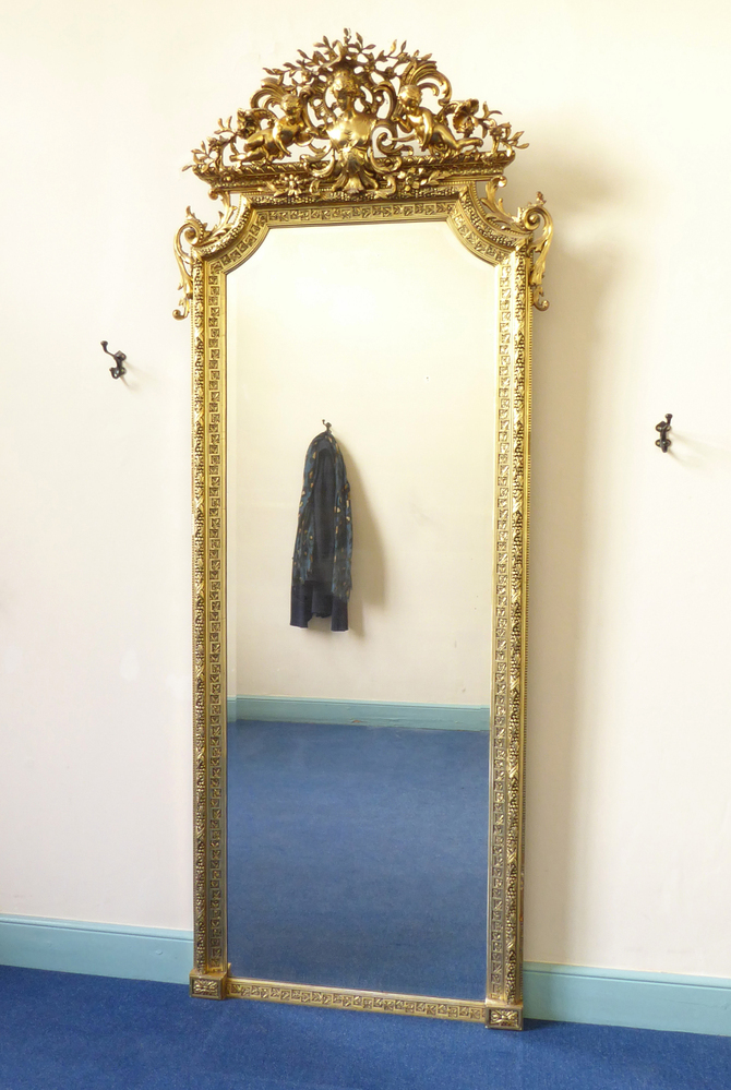 Rechthoekige hoge staande spiegel met kuif, kuif bestaande uit vrouwenbuste te halve lijve met opgestoken haar, vlecht aan achterkant en vijfpuntige stervormige decoratie op borst, geflankeerd door 2 liggende engeltjes zonder vleugels met hoorn, rondom blad- en bloemmotieven, aan linker en rechterbovenkant sierkrul van acanthusblad, facet geslepen glas, onderkant links en rechts rechthoekige houten bewerkte blok met decoratie in het midden, rond buitenrand spiegel blad- en bes- motieven, binnenrand geometrisch zigzaglijn met hulstmotieven, geheel houten achterwand met gips en bladgoud omtrokken. Waarschijnlijk Frankrijk, eind 19e eeuw.