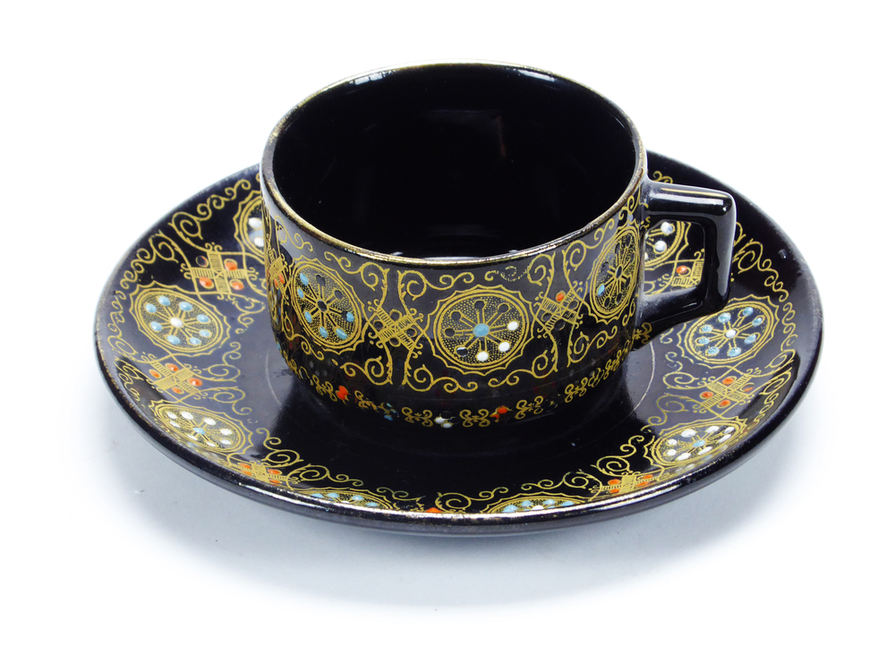 donkerbruin tegen zwart aan aardewerken kop- en schotel, waarschijnlijk gemaakt in Japan, rondom versierd met geometrische goudkleurige motieven , handbeschilderd met emaillen florale motieven, gemerkt aan onderkant met witte emailen stip, afkomstig van zeevarende veenkoloniale familie