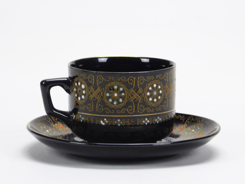 donkerbruin tegen zwart aan aardewerken kop- en schotel, waarschijnlijk gemaakt in Japan, rondom versierd met geometrische goudkleurige motieven , handbeschilderd met emaillen florale motieven, kop gemerkt aan onderkant met 2 lichtblauwe emailen stippen, schotel gemerkt aan onderkant met 3 rode emaillen stippen, afkomstig van zeevarende veenkoloniale familie