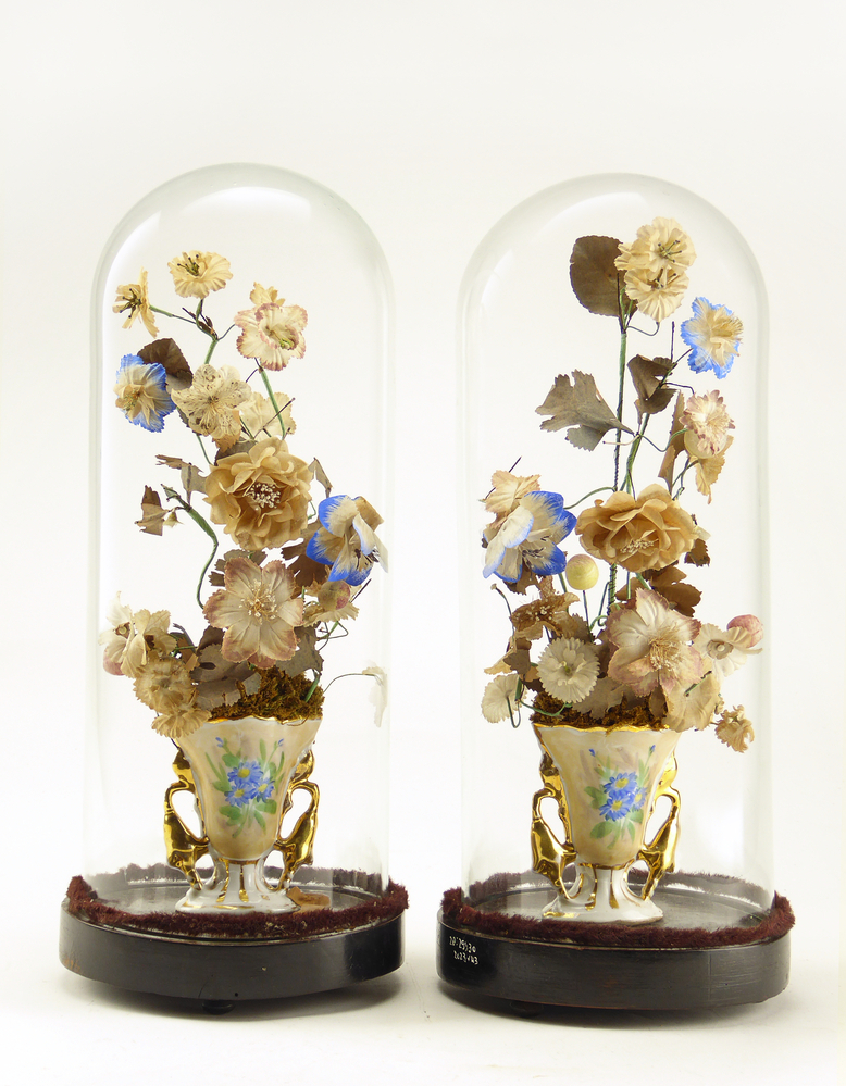 Stolp met kunstbloemen, de stolp is van glas met een bodem van hout met daarin kunstbloemen van textiel, metaal en papier in een wit porceleinen vaasje met goudkleurige versieringen. De rand van de stolp is onderaan omringd door een donkerbruin fluwelen koord. Deze glazen stolpen beschermden kunstbloemen van textiel, papier, ijzerdraad en natuurlijk materiaal in een porseleinen vaas. Tot ver in de twintigste eeuw vormde een glazen stolp met daaronder een bloemstuk, droogboeket, kunstbloemen of het huwelijksboeket een pronkstuk in veel woonkamers. Dergelijke pronkstukken werden onder andere verzameld door Volendammer vissers ter decoratie van hun huis. De glazen stolpen beschermden de tere kostbaarheden tegen stof, beschadiging en uitdroging. Ook fijngevoelige instrumenten zoals klokken en kwetsbare beeldjes werden onder een stolp bewaard. De meeste stolpen zijn inmiddels verdwenen, maar deze kunstbloemen hebben hun stolp behouden wellicht omdat ze erg kwetsbaar zijn., afkomstig van familie Olsder (Meeden, vaderskant) en Meijering (Annerveenschekanaal, moederskant).