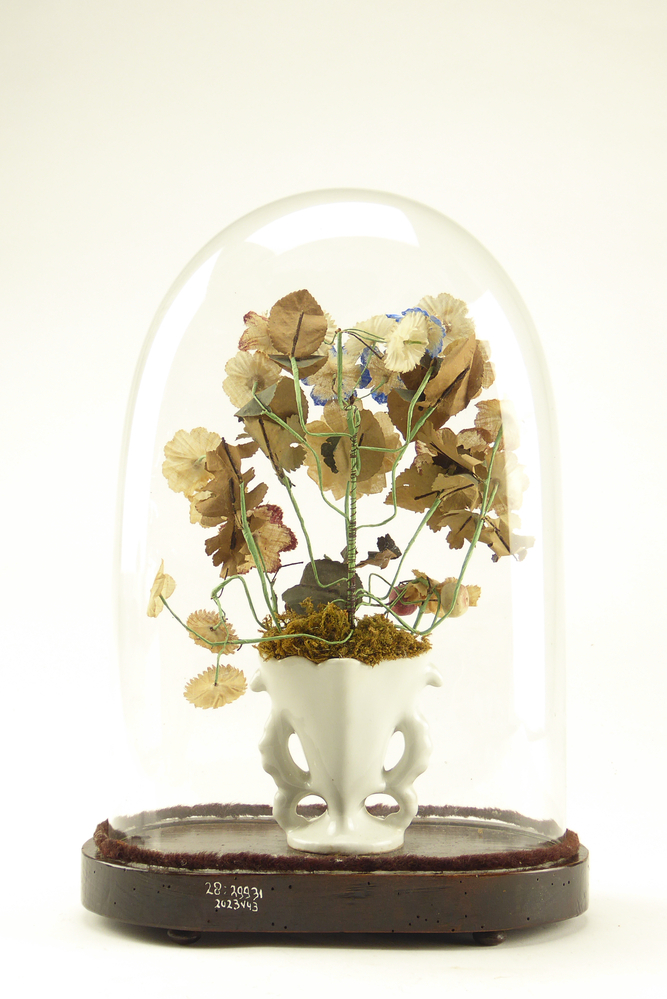 Stolp met kunstbloemen, de stolp is van glas met een bodem van hout met daarin kunstbloemen van textiel, metaal en papier in een wit porceleinen vaasje met goudkleurige versieringen. De rand van de stolp is onderaan omringd door een donkerbruin fluwelen koord. Deze glazen stolpen beschermden kunstbloemen van textiel, papier, ijzerdraad en natuurlijk materiaal in een porseleinen vaas. Tot ver in de twintigste eeuw vormde een glazen stolp met daaronder een bloemstuk, droogboeket, kunstbloemen of het huwelijksboeket een pronkstuk in veel woonkamers. Dergelijke pronkstukken werden onder andere verzameld door Volendammer vissers ter decoratie van hun huis. De glazen stolpen beschermden de tere kostbaarheden tegen stof, beschadiging en uitdroging. Ook fijngevoelige instrumenten zoals klokken en kwetsbare beeldjes werden onder een stolp bewaard. De meeste stolpen zijn inmiddels verdwenen, maar deze kunstbloemen hebben hun stolp behouden wellicht omdat ze erg kwetsbaar zijn., afkomstig van familie Olsder (Meeden, vaderskant) en Meijering (Annerveenschekanaal, moederskant).