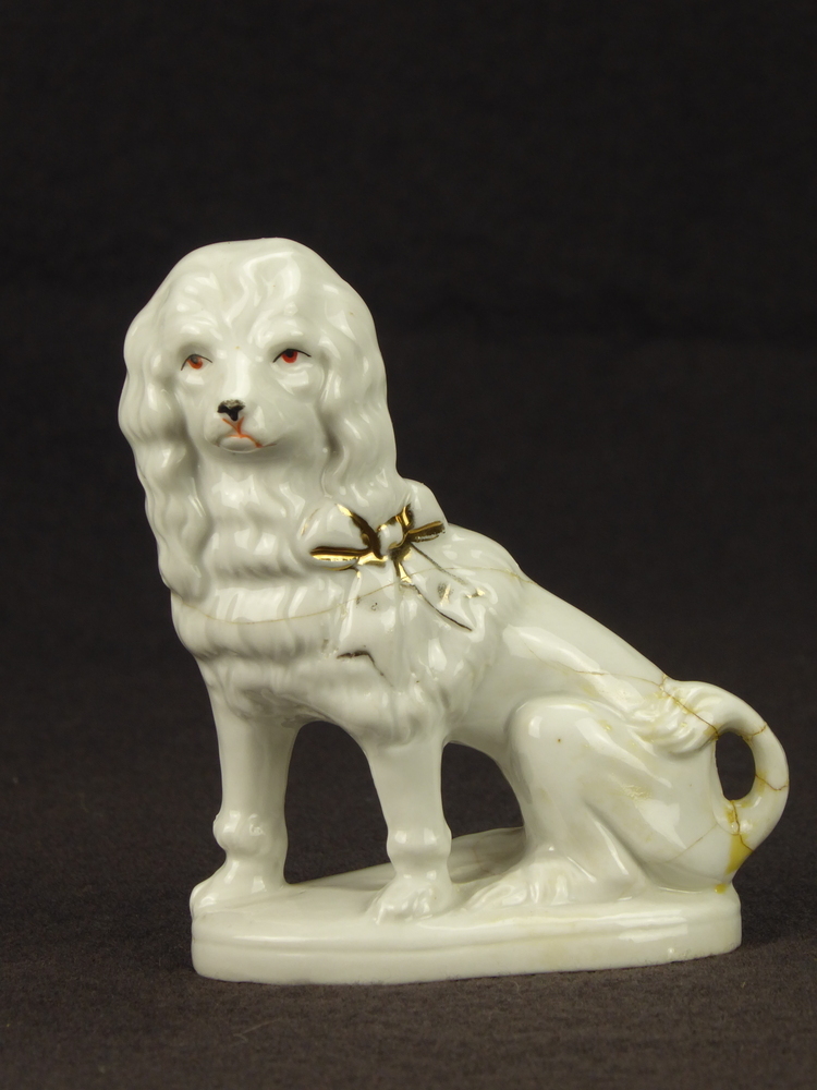 witte steengoed hond, little lion dog, met gouden strik (afgesleten), losse voorpoten, op ovale stander, geheel wit, breuklijnen in lichaam, staart al eens gelijmd, Duits aardewerk naar model van Staffordshire. in rand inscriptie GERMANY en modelnummer 267